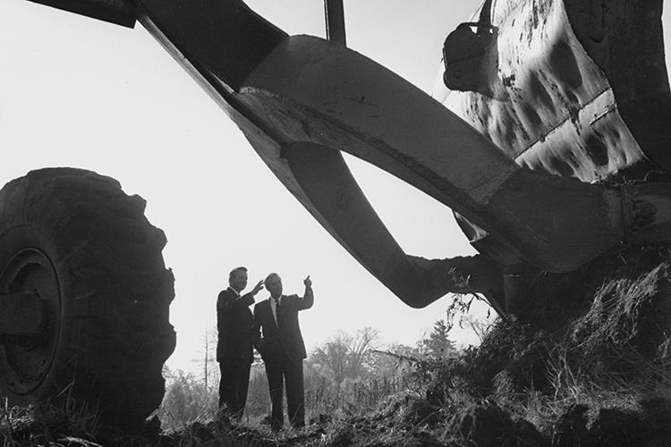 Bill Davis and Carleton Williams are framed under the arch of a backhoe's digging arm.
