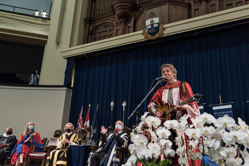Jim Cuddy plays guitar on stage at Convocation Hall while dignitaries clap.