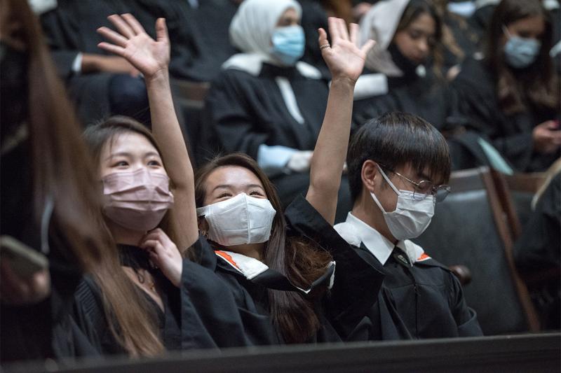 A graduate in robe and masks raises her hands and laughs, sitting in the audience at Convocation Hall.