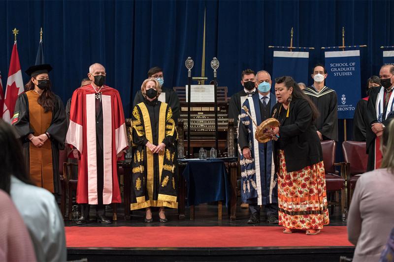 Jenny Blackbird plays a handheld drum on stage at Convocation Hall as dignitaries listen.