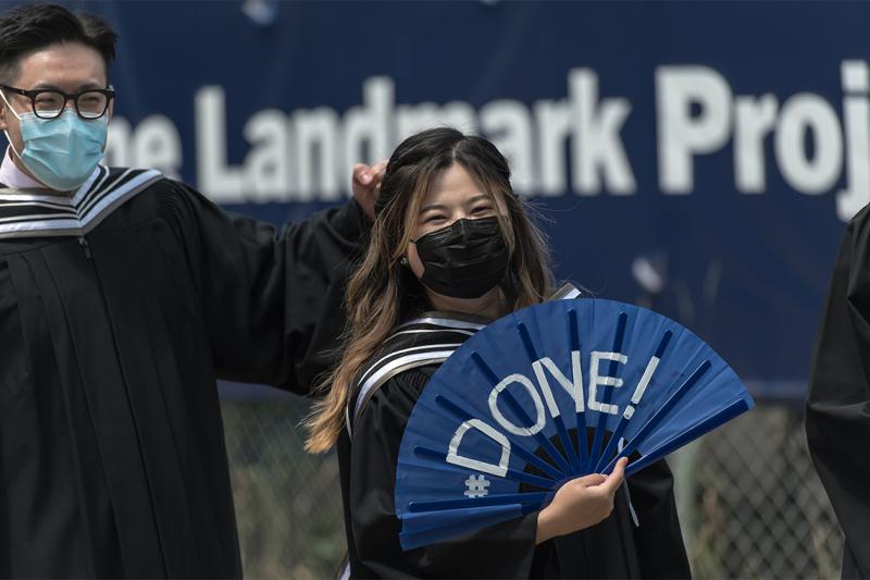 A laughing student in graduation robes and mask waves a paper fan with the text #done.