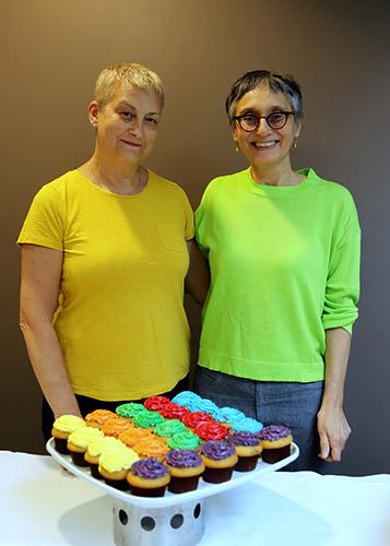 Angela Lange and Amrita Daniere smile, wearing neon T-shirts and standing behind a tray of cupcakes with brightly coloured icing.