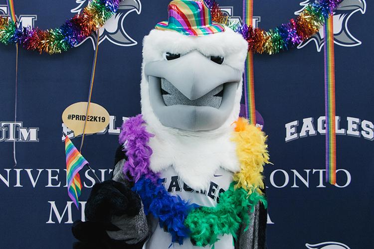 A person in costume wears a giant white eagle head, a shiny rainbow fedora and a rainbow feather garland.