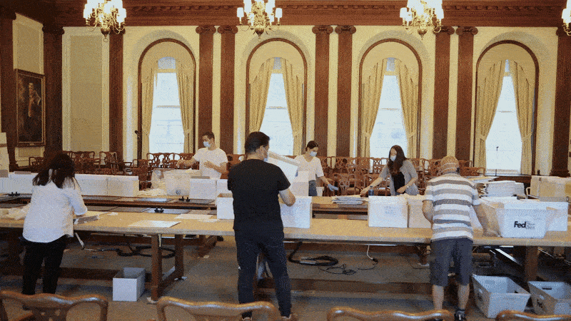 A moving gif image shows people wearing masks and gloves moving quickly to stuff envelopes at a long table.