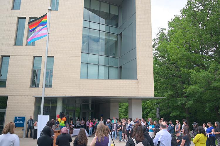 A crowd of people look up at the Pride and Trans flags flying at the top of a flagpole.
