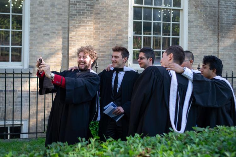 Six young men in academic robes smile as they line up behind a friend holding up a phone.