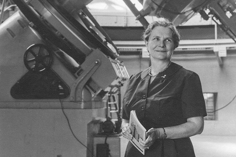 Helen Sawyer Hogg smiles and looks upwards as she stands beneath an enormous telescope.
