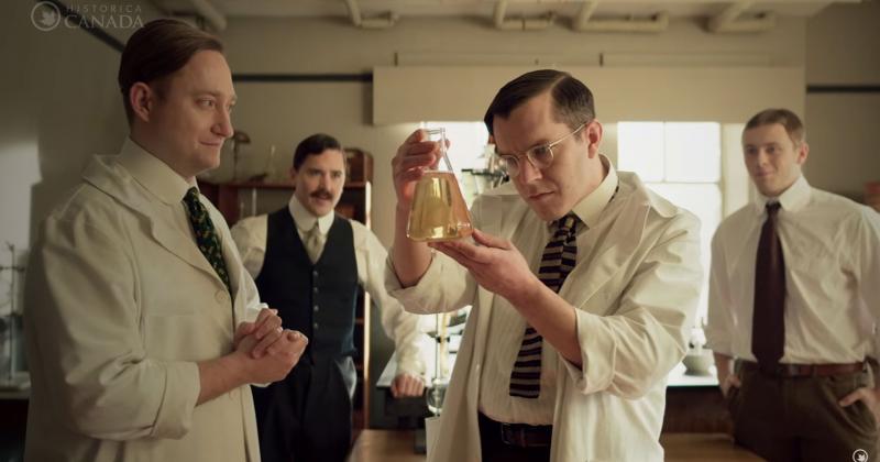 In a screenshot from the Heritage Minute on the discovery of insulin, 4 scientists in 1921 gaze at a beaker of clear liquid.