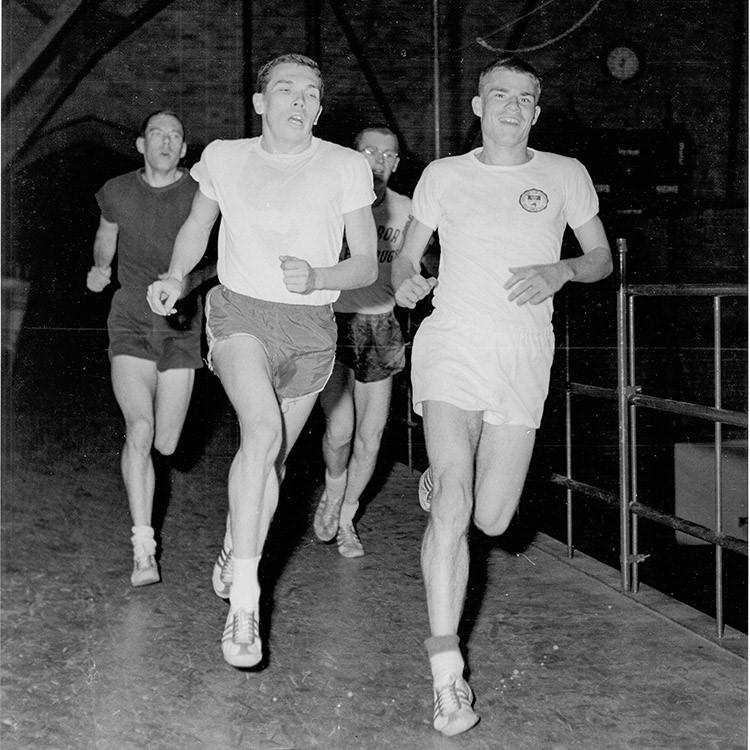 Four young men in shorts and T-shirts race across a gymnasium floor.