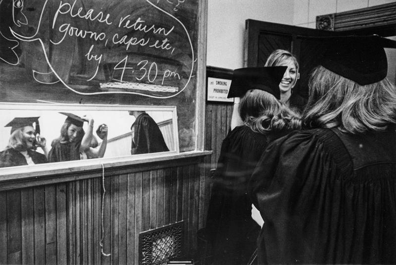 Students laugh as they look in a mirror at themselves wearing mortarboards. On a blackboard: Please return gowns by 4:30 pm.