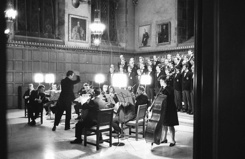 A choir stands on steps behind a dozen musicians at one end of Hart House Great Hall. A conductor gestures energetically.