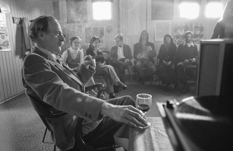 Marshall McLuhan, holding a cigar and a glass of wine, faces a semi-circle of students. The air is very smoky.