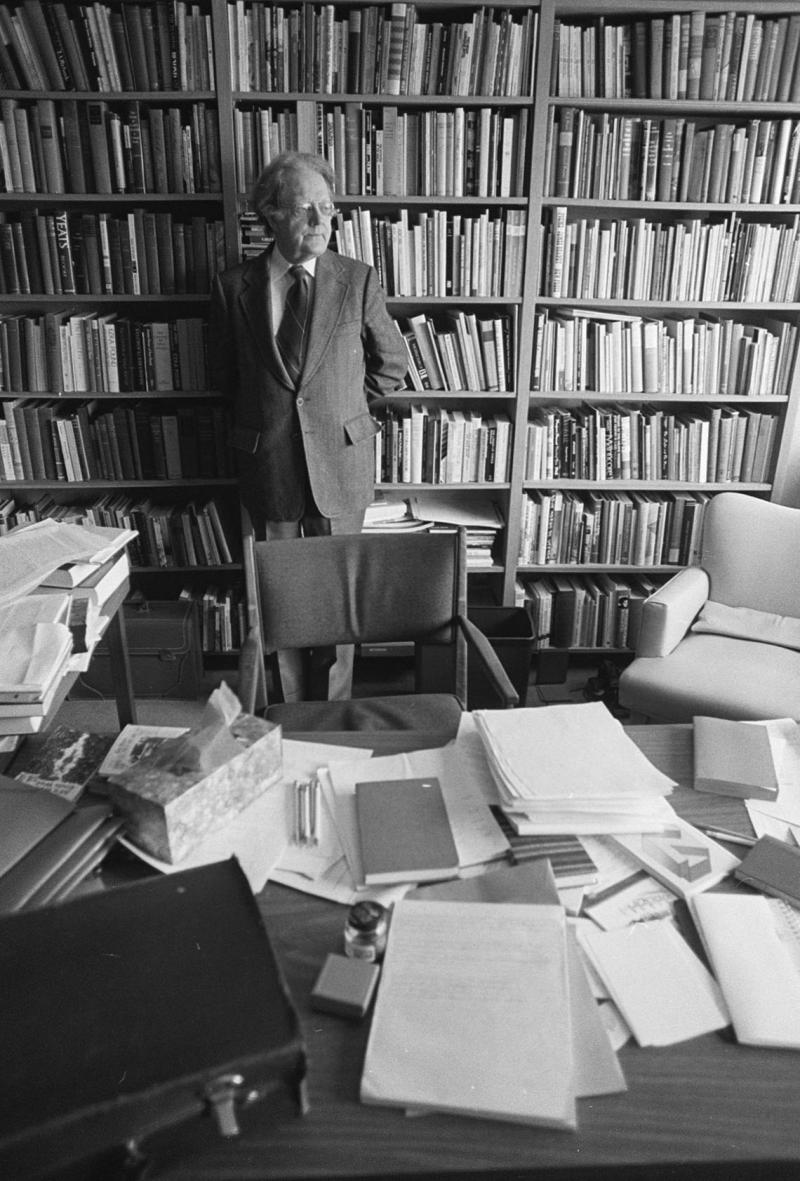 Northrop Frye stands with his back to a huge, crowded bookshelf. In front of him is a desk piled with books and papers.