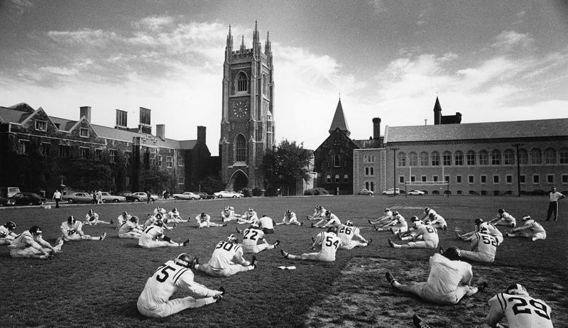 All over the grass of Back Campus, football players sit with legs stretched in a V-shape and touch their toes.