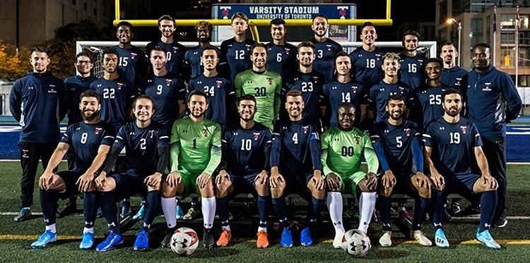 An official soccer team portrait: 28 diverse young men smiling in varsity blues uniforms.