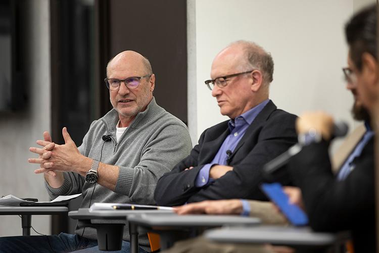 Mark Fox gestures, sitting in a lecture theatre seat next to three other professors who are listening 