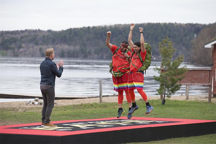 James Makokis and Anthony Johnson jump on a mat by a lakeside, wearing matching orange skirts and bandanas.