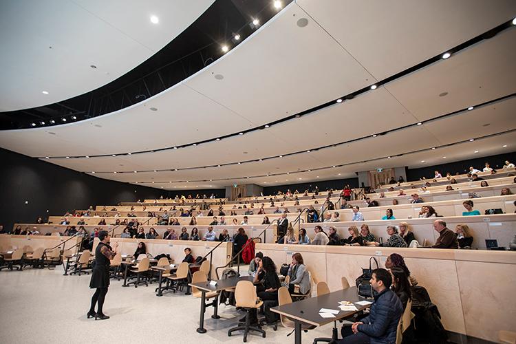 Patricia O'Campo speaks into a microphone and looks up at a full lecture hall, with people sitting at tables  arranged on tiered risers
