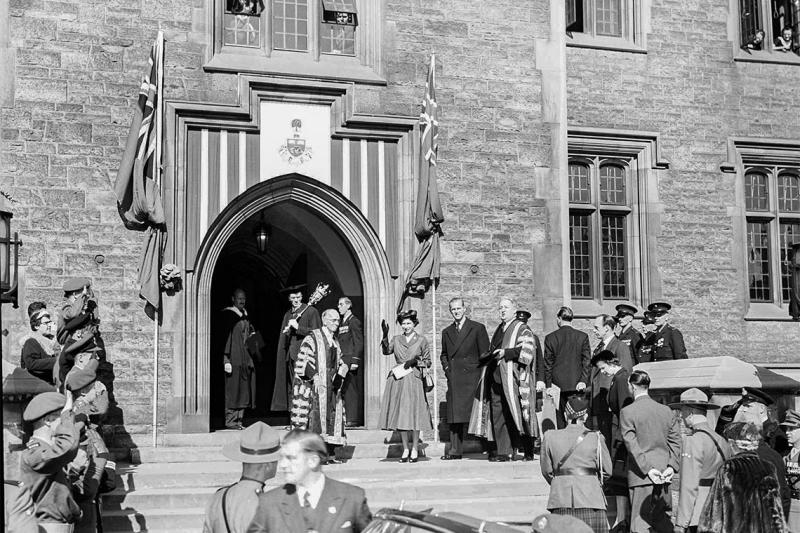 A young Queen Elizabeth II waves as she stands on the steps of Hart House surrounded by dignitaries and saluting soldiers.