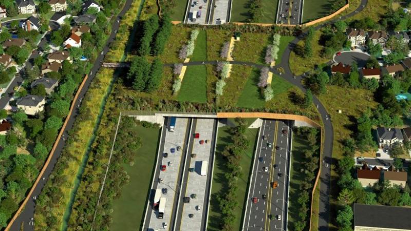 Seen from above, a six-lane highway runs between trees and is crossed by a wide, vegetation-covered, bridge.