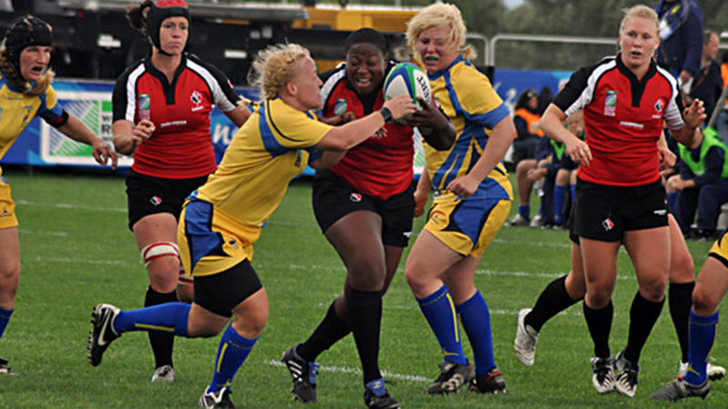 Marlene Donaldson, carrying a rugby ball, ducks between opposing players.