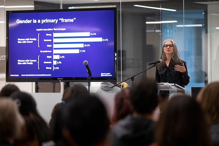 Sarah Kaplan stands at a podium next to a large screen showing a graph and the words "Gender is a primary 'frame'"