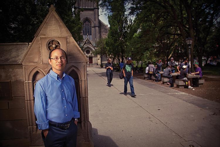 Professor Stephen Hwang standing by a stone wall on a street corner with people walking and sitting at a picnic table in a park in the background (photo by Rob Waymen)