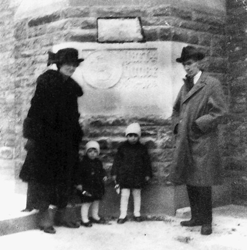 Alice and Vincent Massey stand with two toddlers in front of a carved cornerstone set into a stone wall.