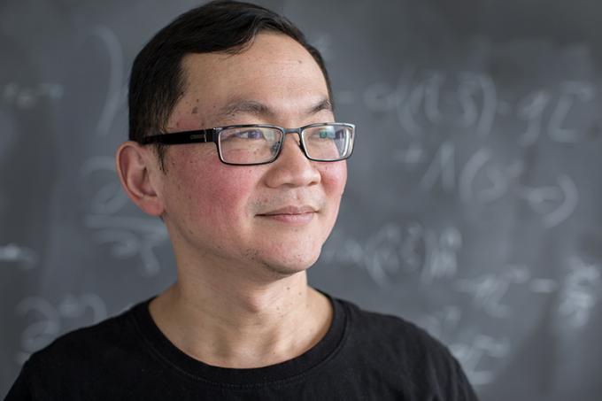 Ue Li stands in front of a blackboard and looks to his left, smiling (photo by Geoffrey Vendeville)