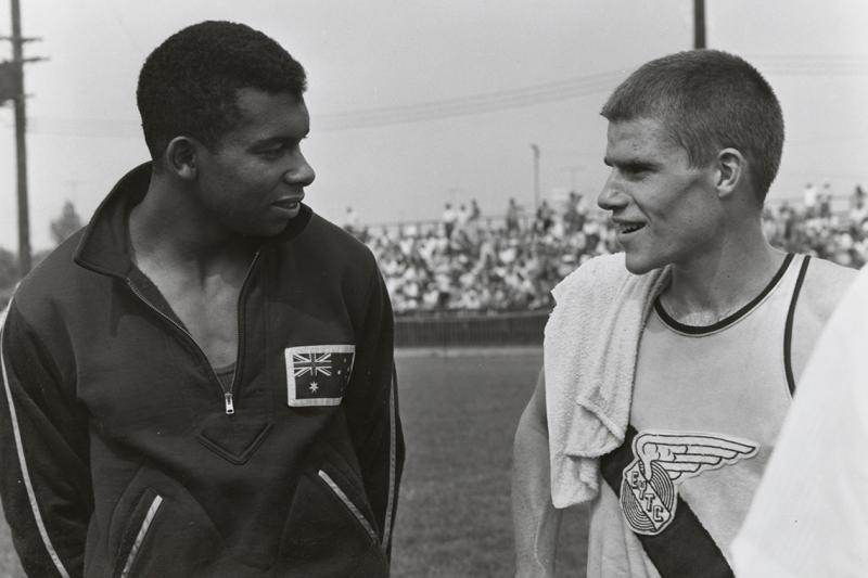 Harry Jerone and Bruce Kidd smile at each other, standing on an athletics field.