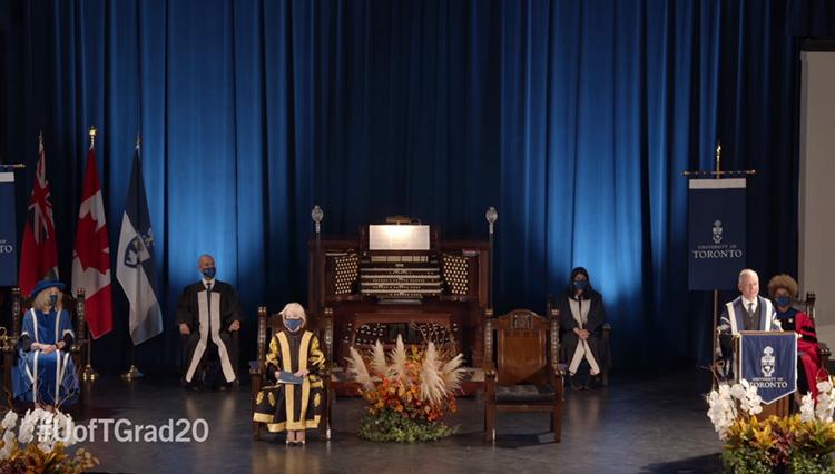 Six officials in academic gowns and masks stand far apart on the stage at Convocation Hall.