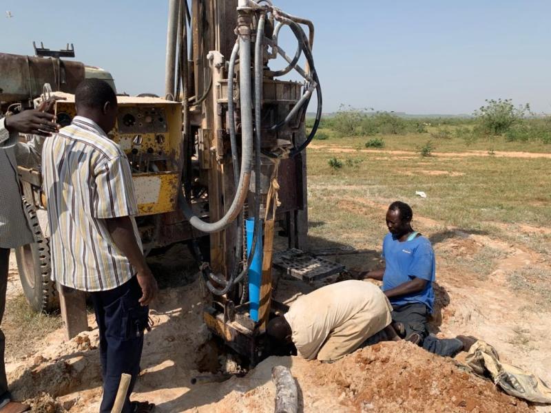 A drilling rig bores a well into a dry field while workers adjust the drill shaft.