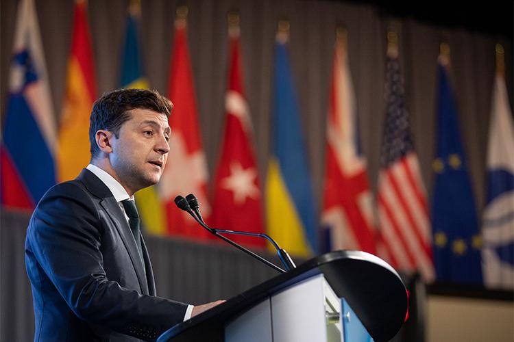 Volodymyr Zelenskyy stands at a podium in front of an array of national flags.