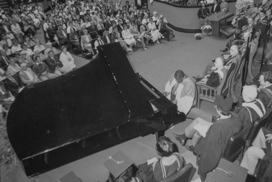 Oscar Peterson plays a grand piano on stage at Convocation Hall, surrounded by professors in academic robes.