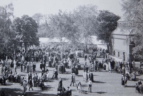 In June 1926, hundreds of people in formal clothes and hats chat in groups on a lawn by a tent and trees.