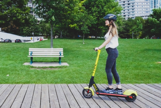 A woman wearing a helmet stands on a Roll brand e-scooter as she rolls along a boardwalk past a park bench.