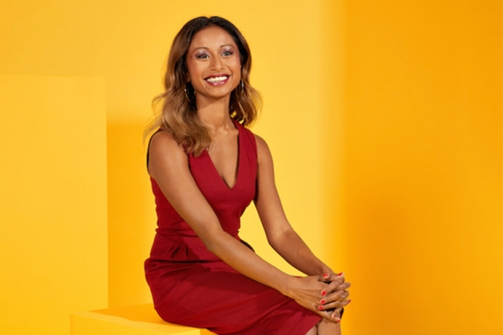 On this a page from Precedent magazine, Janani Shanmuganathan sits on a cube against a sun-coloured background, smiling.