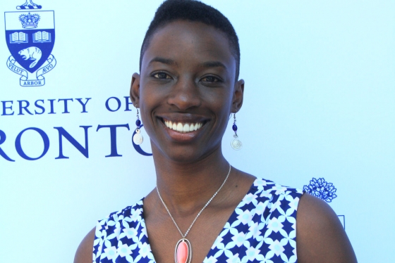 Janelle Joseph smiles while standing in front of a wall with the University of Toronto logo.