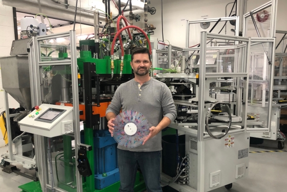 Rob Brown holds a vinyl disc with colourful markings. Behind him is a large press machine that nearly fills the room.