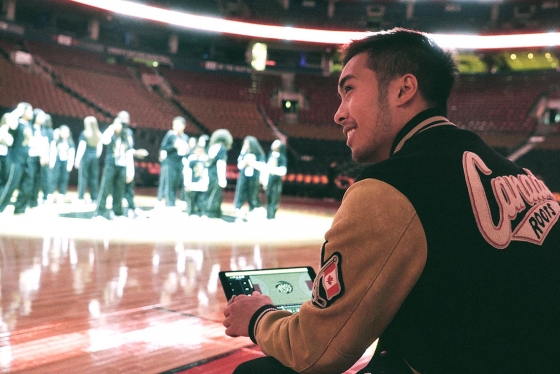 Axel Villamil sits in the foreground holding a tablet with his app and watching the Raptors dancers gather on the court in the background (photo courtesy of StageKeep) 