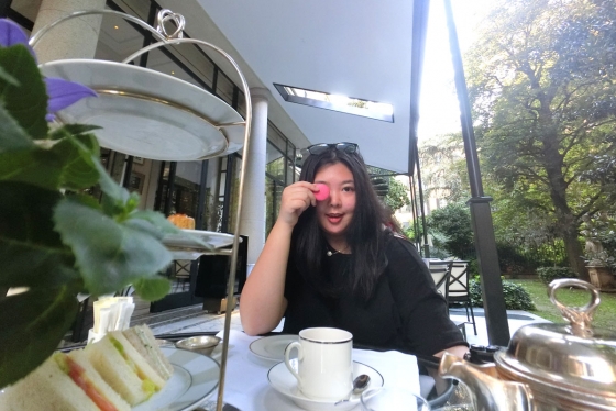 Joanna Luo smiles and holds a pink macaron cookie over one eye, while sitting at an outdoor cafe.