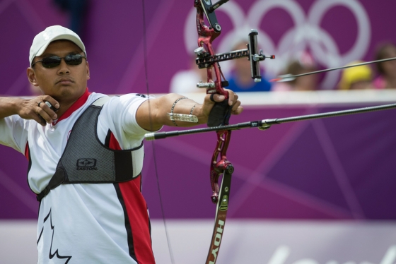 Crispin Duenas releases an arrow from his bow. The Olympic rings are seen on a wall in the distance.