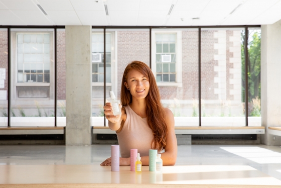 Laura Burget smiles and holds out a small bottle of cosmetics, sitting behind a desk with more products standing on it.