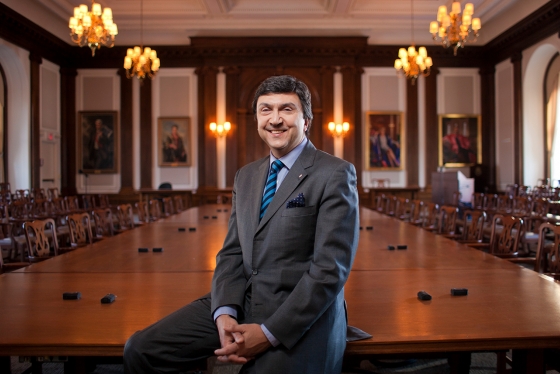 David Naylor smiles while leaning on a  polished conference table in a large boardroom, decorated with painted portraits and old-fashioned chairs.