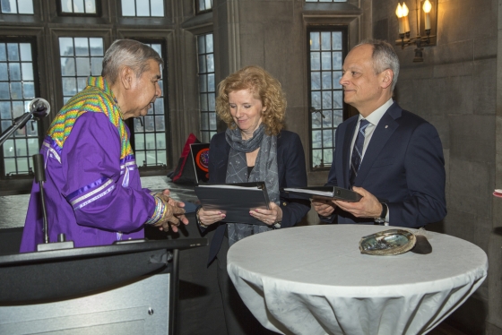 Elder Andrew Wesley, wearing formal Indigenous regalia, hands binders to Cheryl Regehr and Meric Gertler by a table holding a sacred abalone shell (photo by Johnny Guatto)