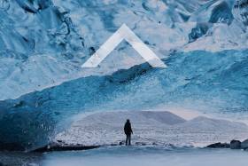 Man stands underneath an ice bridge looking at a mountain in the distance. A caret icon in the middle of the image.