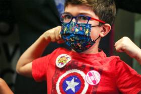 A young boy flexes his muscles. He's wearing a mask, a superhero t-shirt, and a sticker that says I got my COVID-19 vaccine.