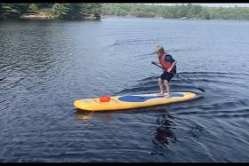 On a lake, a boy stands and jumps on the stern of a paddleboard. Ripples behind show he's moving forward.