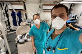 Sgt. Angela Brownell and Jesse Barker, wearing scrubs and masks, stand beside a dental chair inside a large tent.