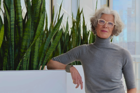 A woman in a grey turtleneck and glasses poses next to some potted plants